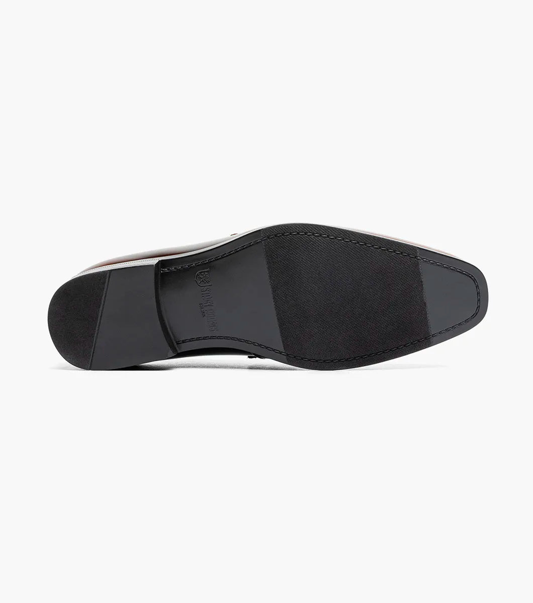 The Stacy Adams Spratley Moc Toe Saddle Slip On (Brown, 25686-200) is a men’s loafer with a glossy leather finish, cushioned insole, and velvet strap detail, displayed angled on a plain white background.
