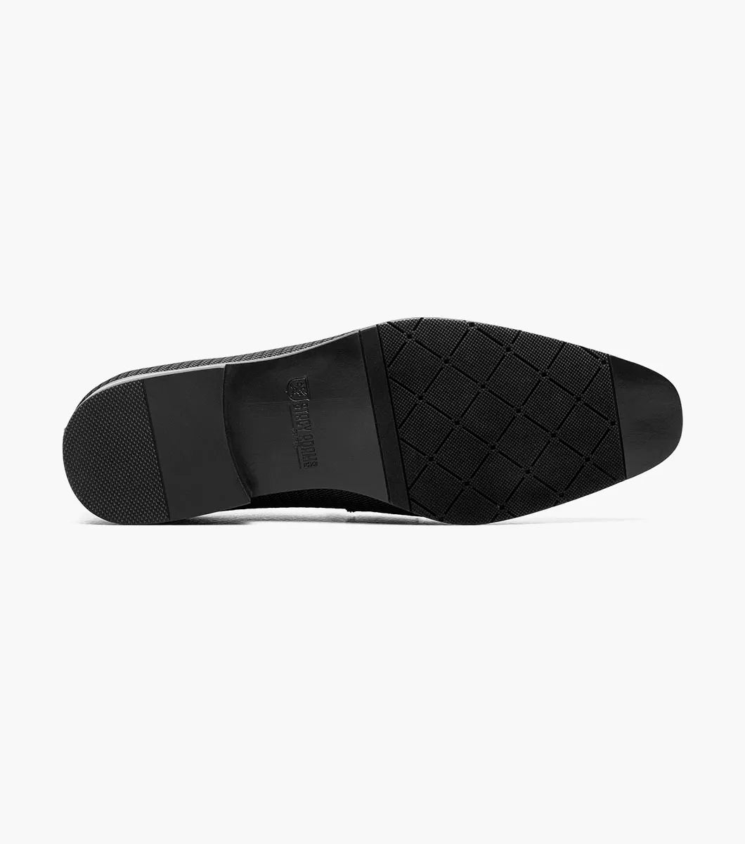 The STACY ADAMS Tazzi Plain Toe Saddle Ornament Slip On (25529-001) is a black textured men’s loafer with leather linings, Memory Foam footbed, decorative saddle band and emblem, and low heel—shown on a plain white background.