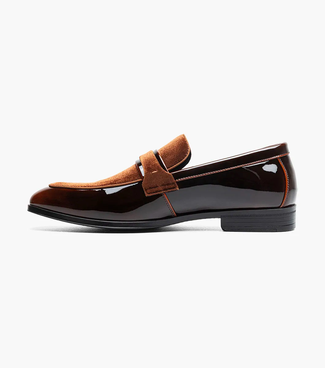 The Stacy Adams Spratley Moc Toe Saddle Slip On (Brown, 25686-200) is a men’s loafer with a glossy leather finish, cushioned insole, and velvet strap detail, displayed angled on a plain white background.