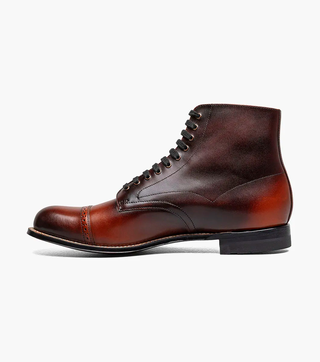 The Stacy Adams Madison Cap Toe Lace Up Boot in Cognac (00110-221) features brown leather, black laces, brogue detailing, and a stacked heel. This stylish STACY ADAMS boot is shown on a white background.