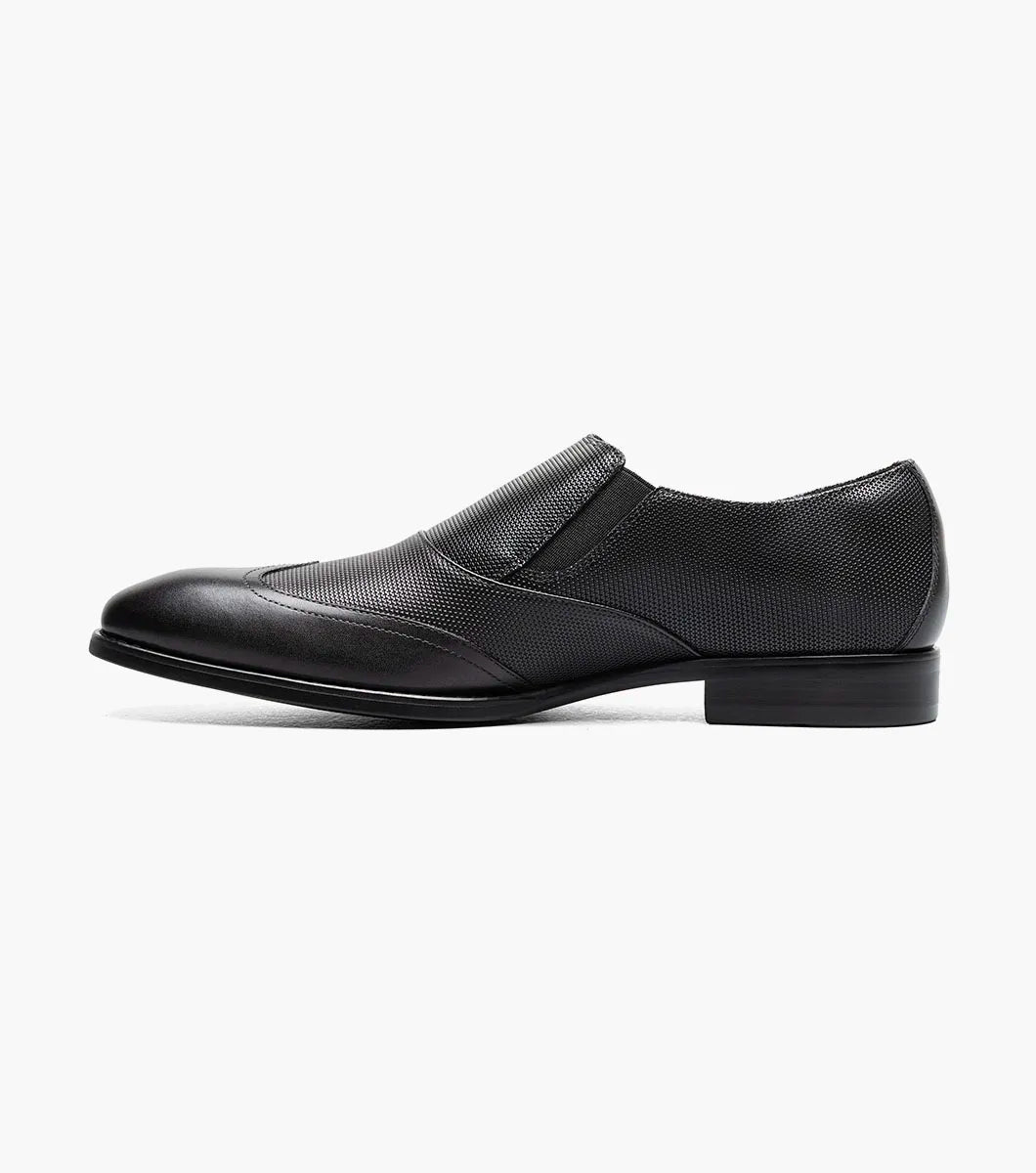 The STACY ADAMS Lockwood Wingtip Ornament Slip On in black (25767-001) features a textured upper, wingtip design, and is displayed on a white background.