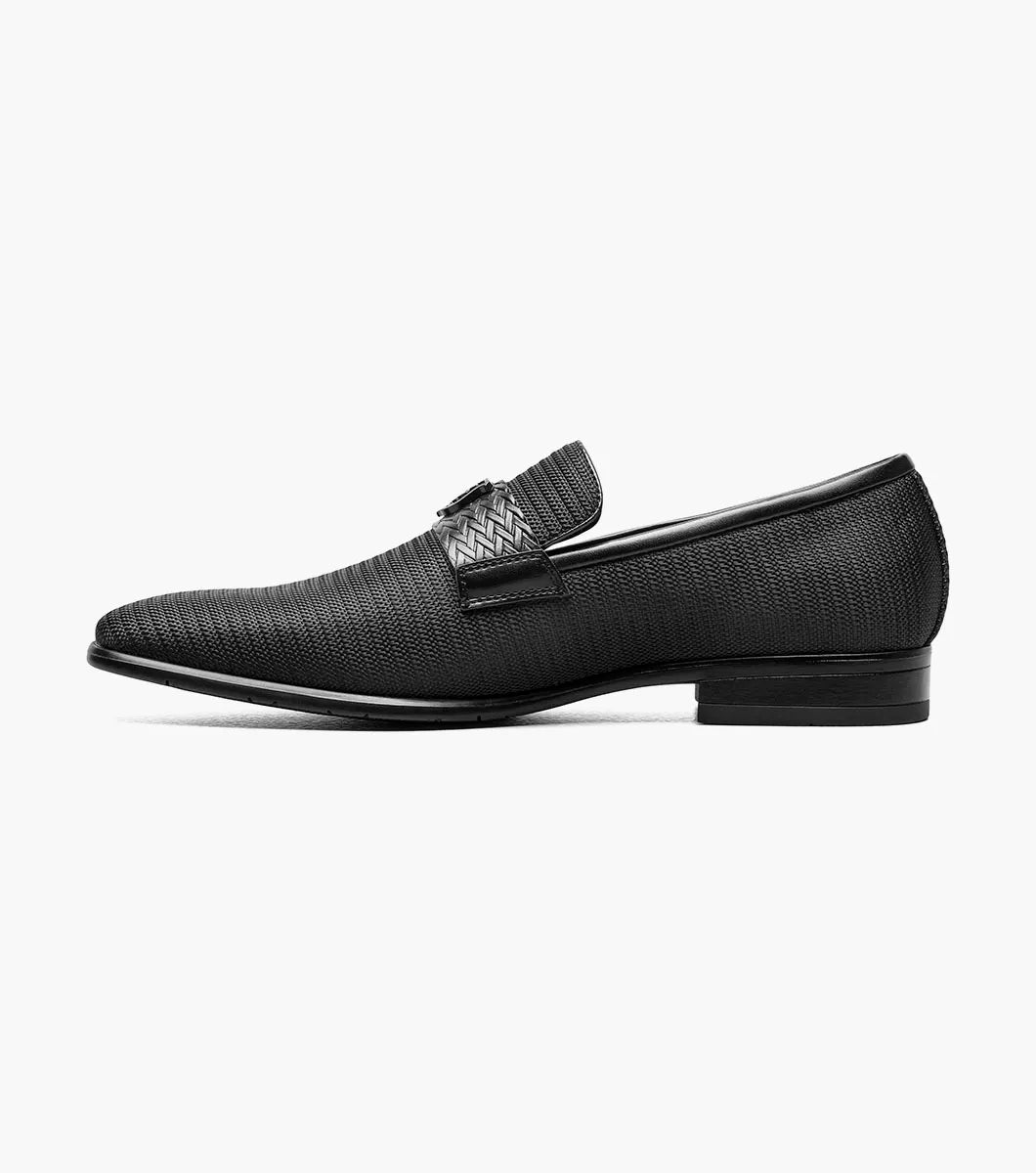 The STACY ADAMS Tazzi Plain Toe Saddle Ornament Slip On (25529-001) is a black textured men’s loafer with leather linings, Memory Foam footbed, decorative saddle band and emblem, and low heel—shown on a plain white background.