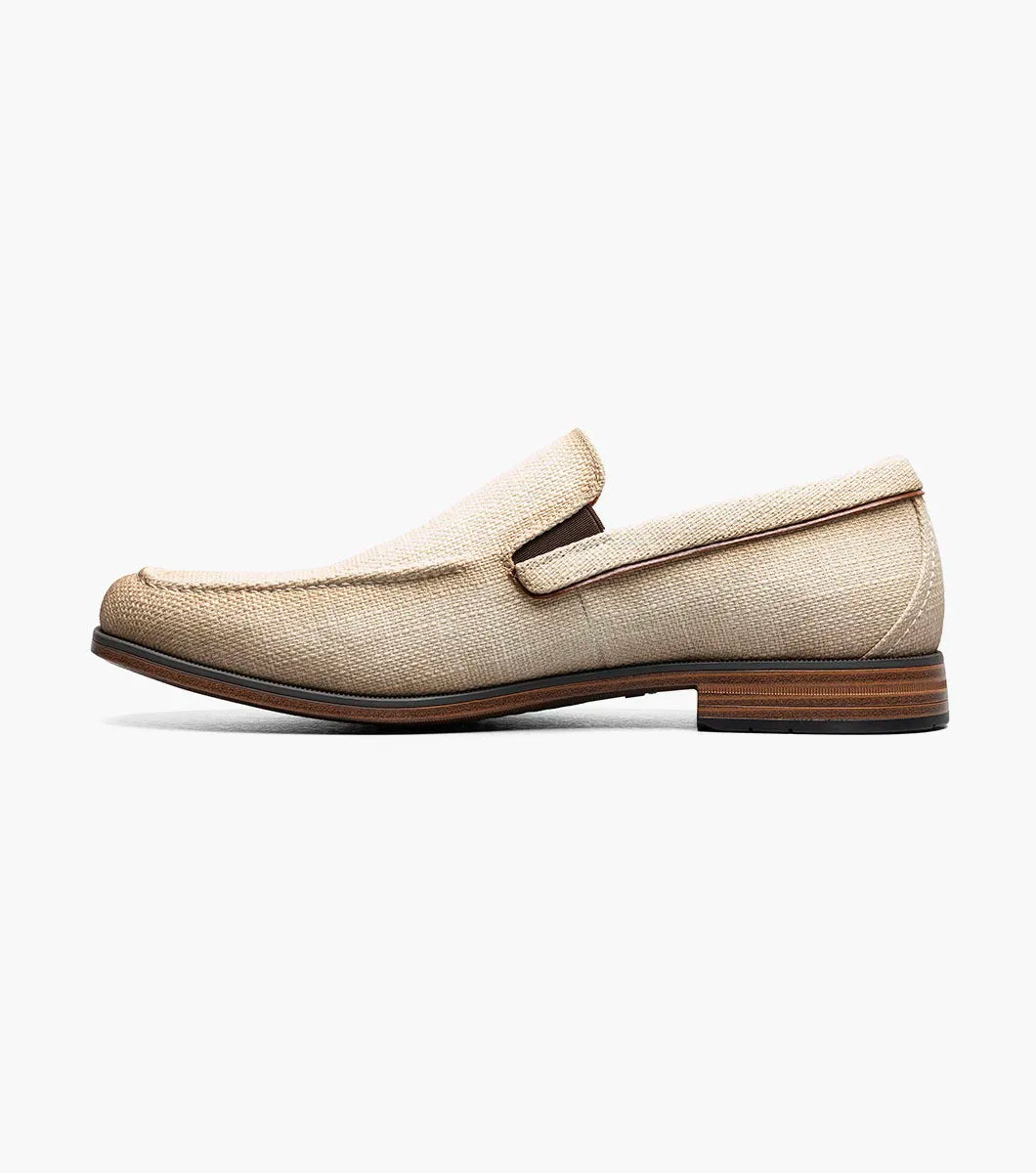 The Stacy Adams Ridgely Moc Toe Slip On (Natural) features a textured fabric upper, brown trim, anatomical arch support, and a low wooden heel for comfort, shown here on a white background.