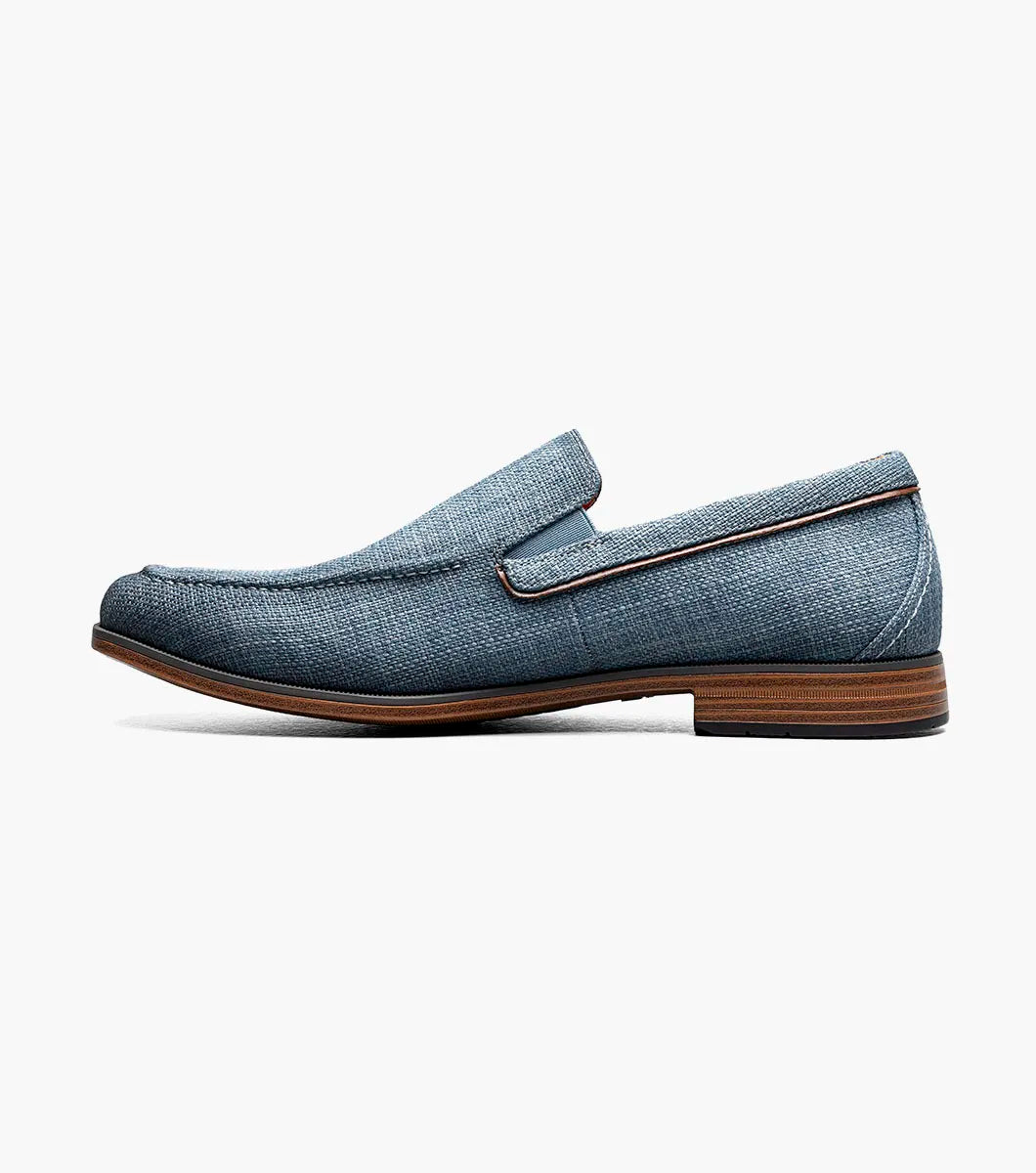 The Stacy Adams Ridgely Moc Toe Slip On in navy features a textured fabric upper, breathable leather lining, brown trim, and a low stacked wooden heel, shown against a white background.