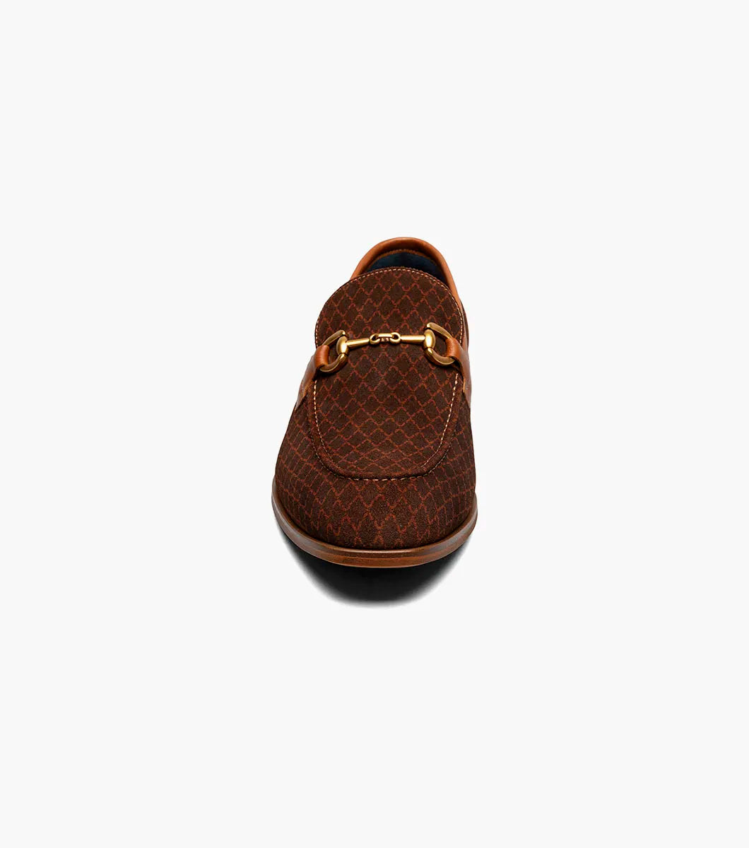 The Stacy Adams Brinkley Moc Toe Bit Slip On in tan features a patterned fabric upper, leather trim, gold metal bit ornament, and a low wooden heel, displayed on a white background.