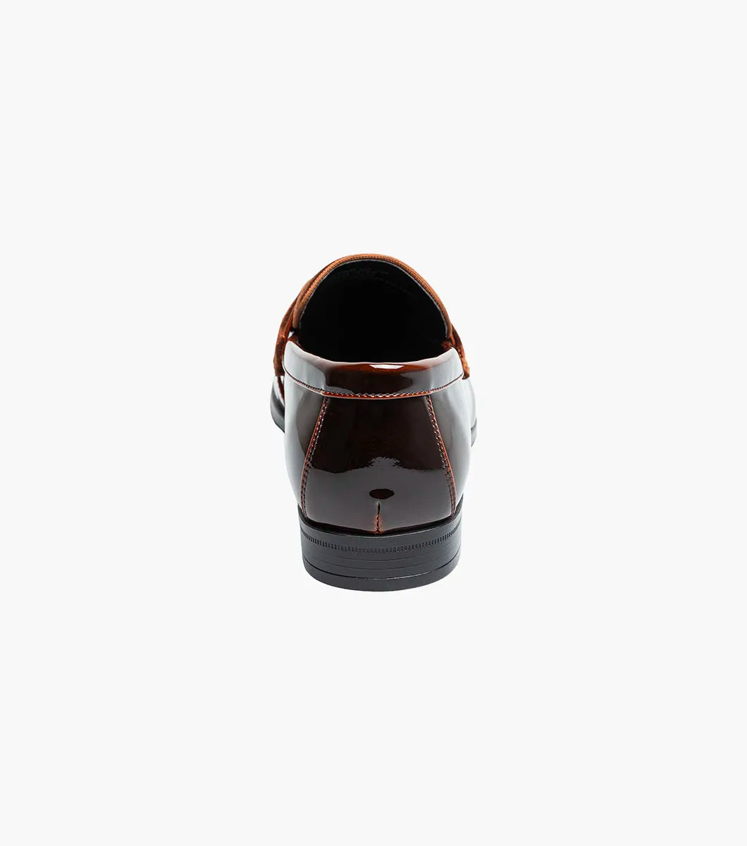 The Stacy Adams Spratley Moc Toe Saddle Slip On (Brown, 25686-200) is a men’s loafer with a glossy leather finish, cushioned insole, and velvet strap detail, displayed angled on a plain white background.
