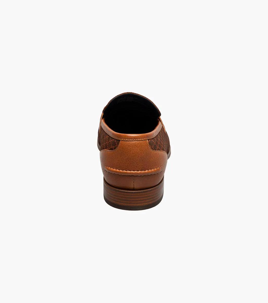 The Stacy Adams Brinkley Moc Toe Bit Slip On in tan features a patterned fabric upper, leather trim, gold metal bit ornament, and a low wooden heel, displayed on a white background.