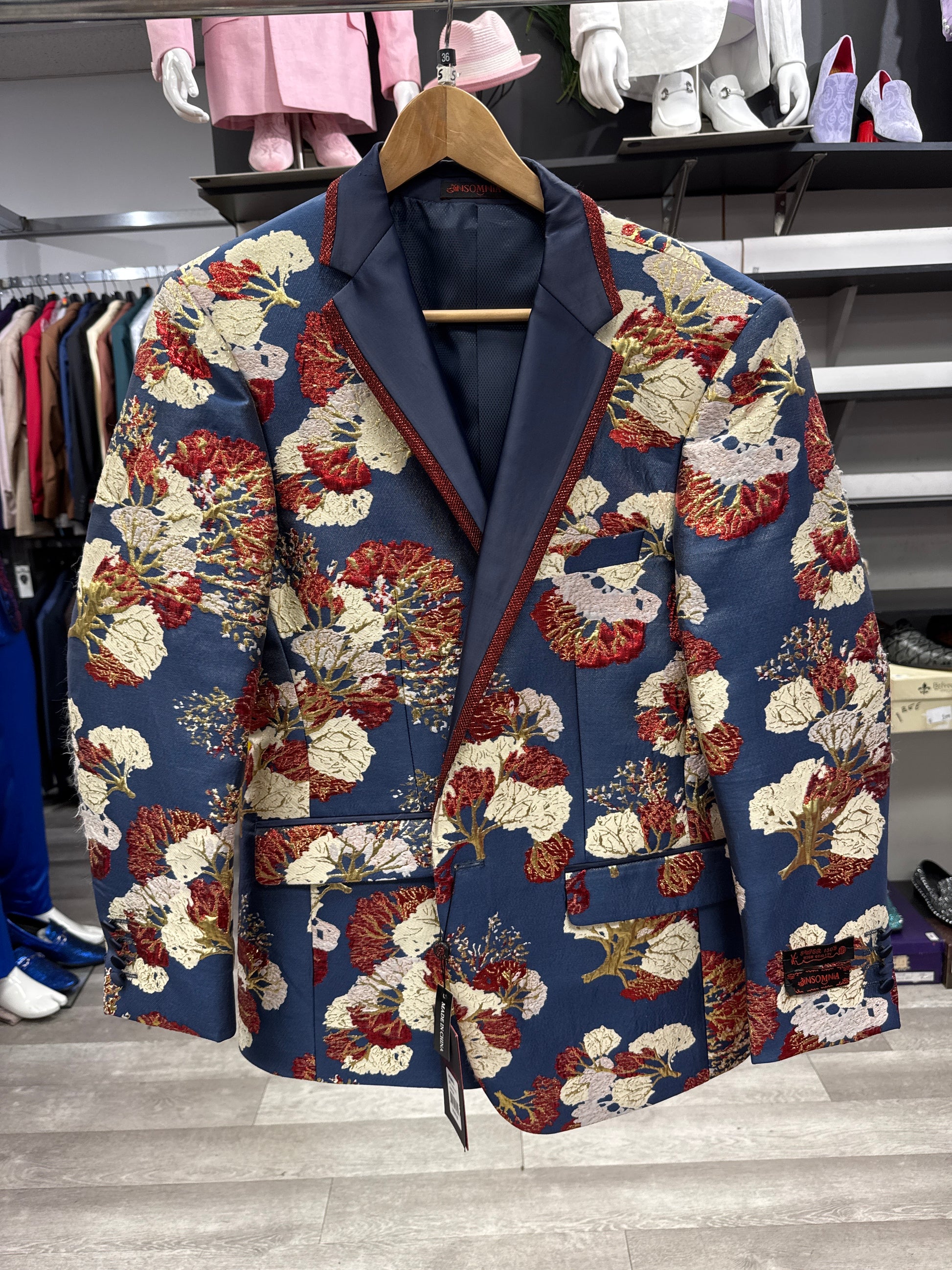 The EJ SAMUEL JACKET 17 CLEARANCE, featuring a blue base with a red, beige, and gold floral pattern, hangs on a wooden hanger in the clothing store. This item is final sale.
