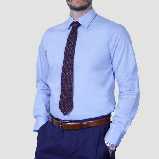 A person wearing an ARTURO Modern Fit Long Sleeve Light Blue Dress Shirt, a dark patterned tie, and a brown belt, with hands in pants pockets.