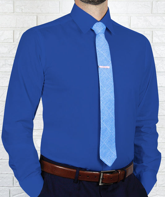 An individual wearing an Arturo Slim Fit Long Sleeve Royal Blue Dress Shirt, a light blue tie, and a brown belt is standing against a white brick wall.