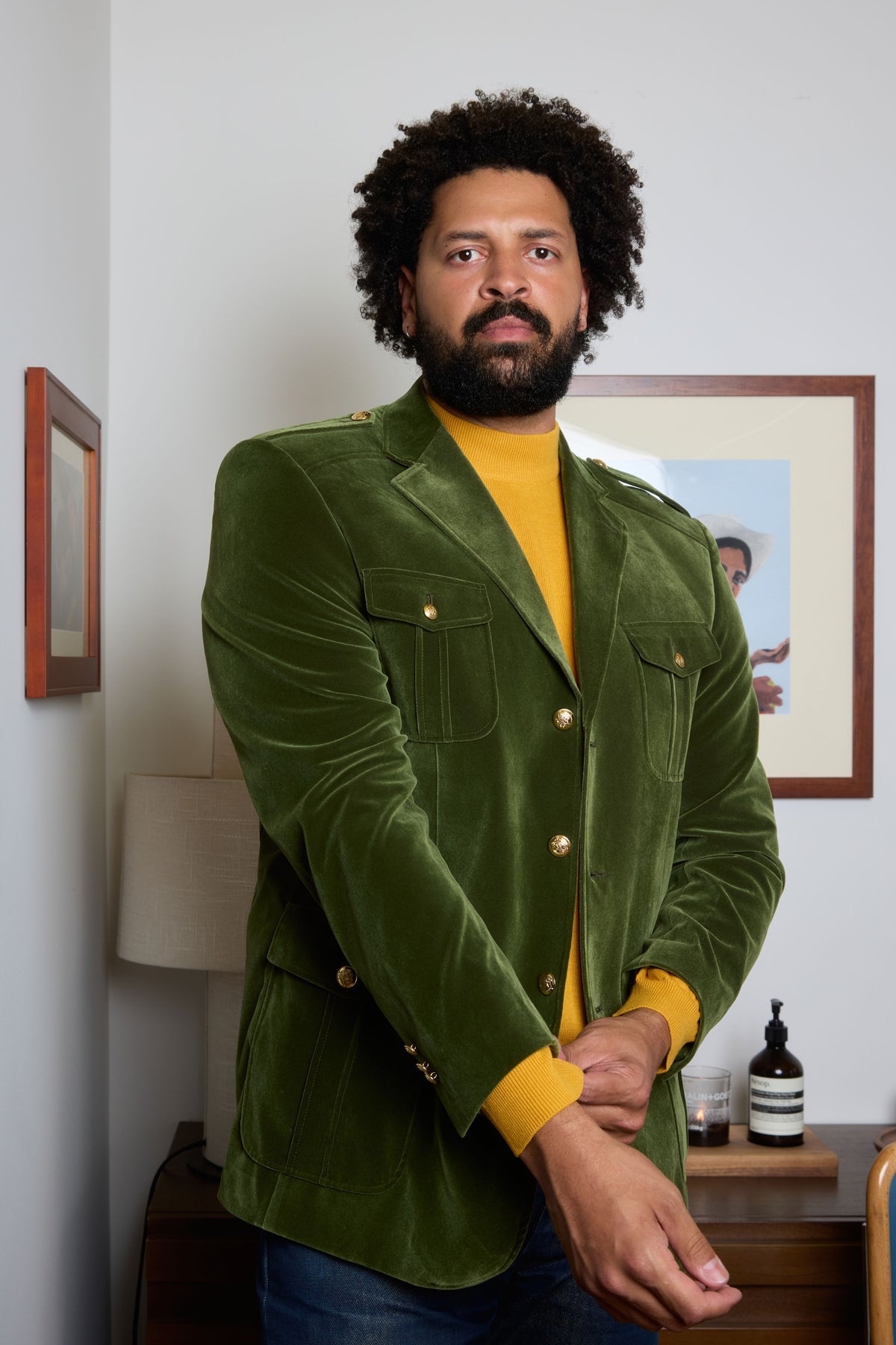 A man wears the Inserch Velvet Military Blazer BL569-19 Olive by Inserch over a blue turtleneck and blue jeans, standing against a beige background.