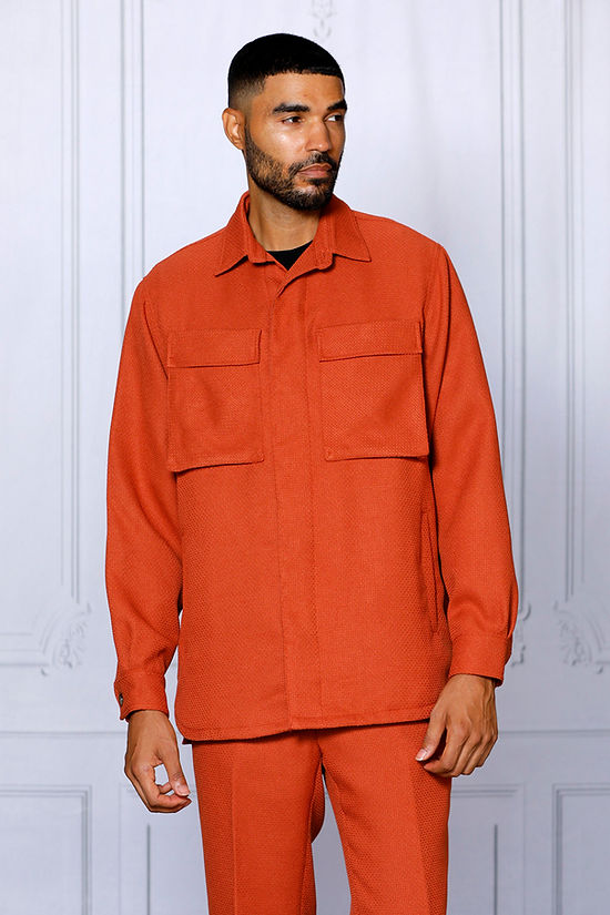A man wearing the INSOMNIA CAVALLI 2PC Coral Leisure Suit by Insomnia, featuring a textured button-up shirt with chest pockets and matching pants, stands against a light-colored paneled wall.