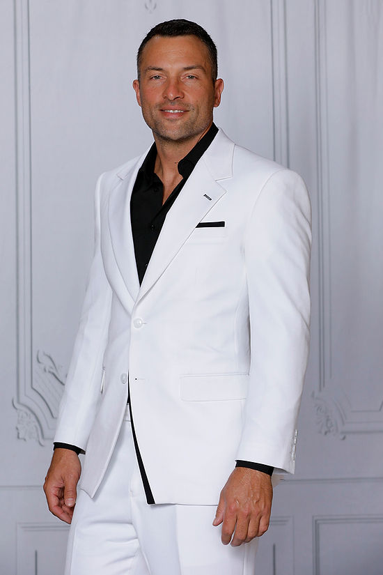 A man wears the INSOMNIA PURPLE LABEL 2PC Tailored Fit White Black 4 Way Stretch Spandex Suit by Insomnia with a black dress shirt, standing before a light paneled wall and looking at the camera.