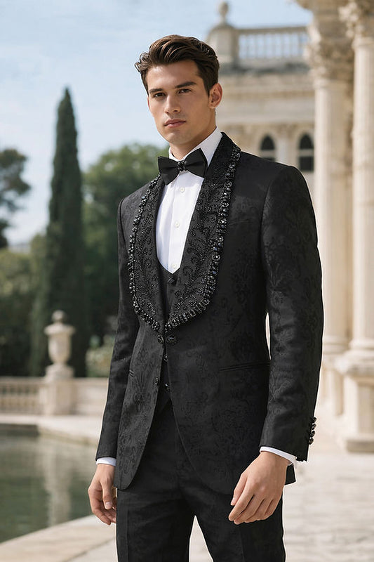 A man wears the MANZINI ASPEN 3-Piece Slim-Fit Men's Fancy Rhinestone Woven Pattern Tuxedo in black by Manzini, with a white shirt and black bow tie, standing outdoors near a classical building and reflecting pool, ready for a formal event.