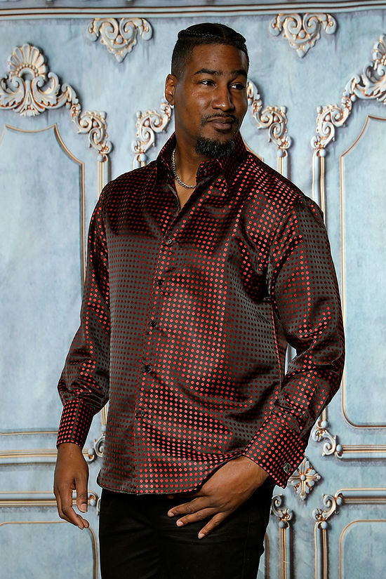 A man wearing a Manzini MZT-164 Small Polka-Dot Long Sleeve Satin Shirt in black with red dots stands before an ornate, light blue decorative wall.