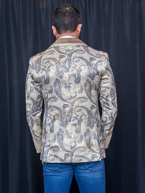 A man wearing the Manzini MANZINI MZS-566 Men's Tailored-Fit Fancy Velvet Paisley Tuxedo Dinner Jacket in brown, paired with beige lapels, a beige bow tie, white shirt, and blue jeans stands before a plain black curtain.