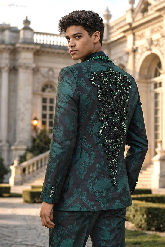 A man wearing the Manzini Aspen 3-Piece Slim-Fit Men's Fancy Rhinestone Woven Pattern Tuxedo in hunter green and sunglasses stands on a cobblestone street lined with buildings.