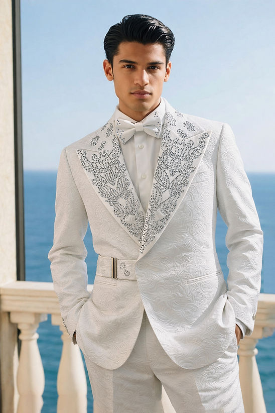 A man in a Manzini MANZINI PREMIUM Men's 2PC Double Breast Slim-Fit Shiny Rhinestone Floral Pattern Suit in white stands on a balcony overlooking the ocean, his elegant suit featuring jeweled details and a floral texture.