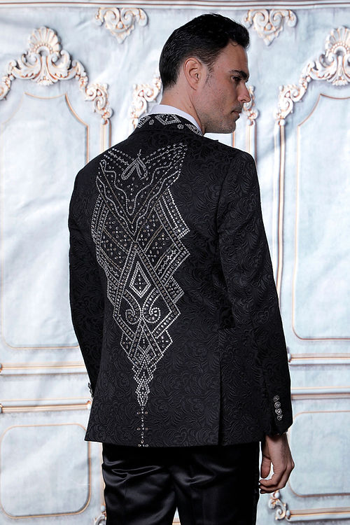 A man in a MANZINI MZS-603 jacket featuring a tailored fit, shiny rhinestones, and a black/silver floral pattern, stands before an ornate light-colored wall.