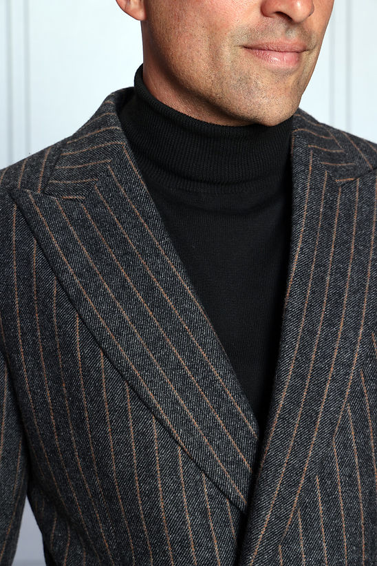 A man stands against a light-paneled background, wearing the Manzini MANZINI MZW-702 Men's Pinstripe Blazer Coat with Hook Belt in charcoal over a black turtleneck and black pants.