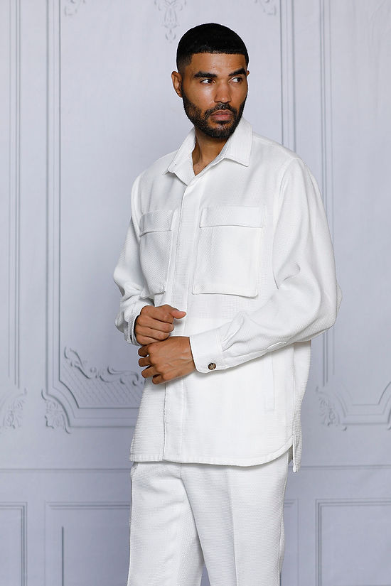 A man stands against a light-paneled wall wearing the INSOMNIA CAVALLI 2PC White Leisure Suit by Insomnia, featuring two large chest pockets and matching white pants—a refined take on casual attire.