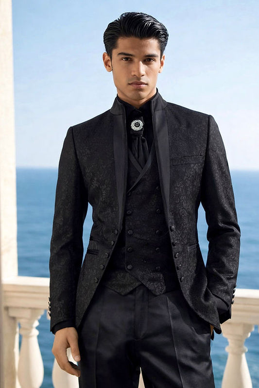 A man stands on a balcony overlooking the sea, wearing the Manzini OAK 3-Piece Slim-Fit Baroque Gothic Tuxedo with satin trim in black, paired with a high-collared shirt and a decorative brooch.