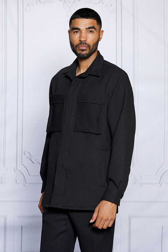 A man with short hair and a beard stands against a white decorative wall, wearing the Insomnia INSOMNIA ROBERTO 2PC Black Leisure Suit made from a soft poly rayon blend.