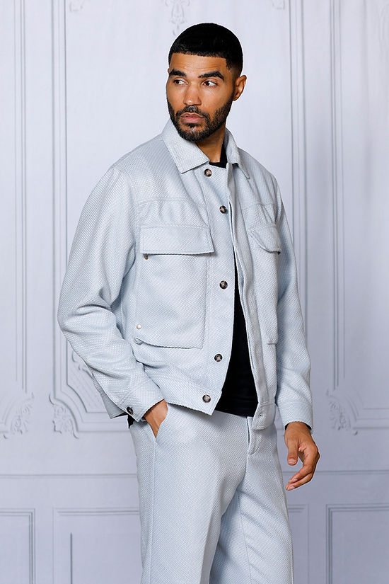 A man wearing the INSOMNIA FENDI 2PC Silver Leisure Suit by Insomnia stands against a decorative light-colored wall, looking to his left.