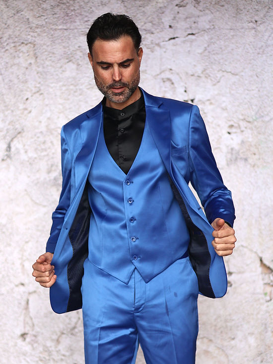 A man wears the INSOMNIA CLASSICO 3PC Tailored Fit Satin Spandex Royal Suit by Insomnia with a black shirt, standing against a textured light wall—ideal for a stylish night out.