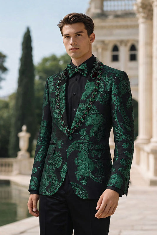 A man stands outdoors wearing the MANZINI MZS-602 Men's Rhinestone Shiny Metallic Floral Tuxedo Prom Jacket in hunter green by Manzini, paired with a matching bow tie, in front of a classical building and greenery.