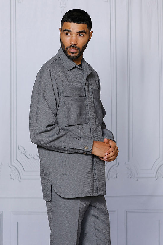 A man poses indoors against a light decorative wall, wearing the INSOMNIA CAVALLI 2PC Charcoal Leisure Suit by Insomnia—a matching poly-rayon blend suit with a textured button-up shirt with two pockets and coordinating grey pants.