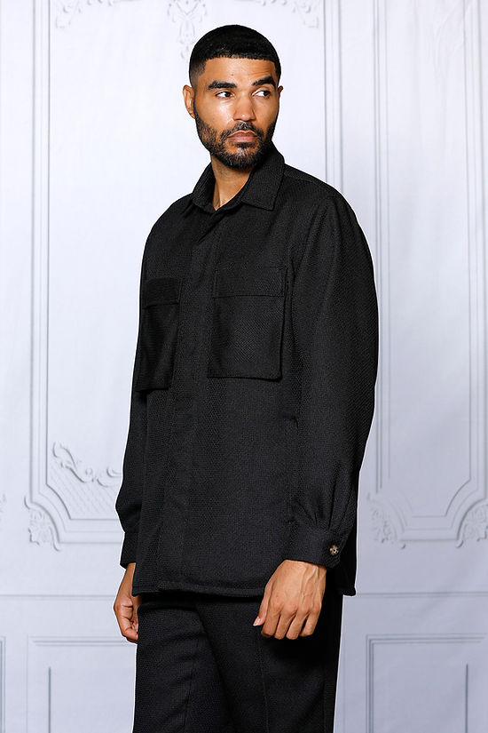 A man faces forward in the Insomnia Cavalli 2PC Black Leisure Suit by Insomnia, featuring a black button-up shirt with front pockets and matching pants, set against a light-colored, ornate wall background.