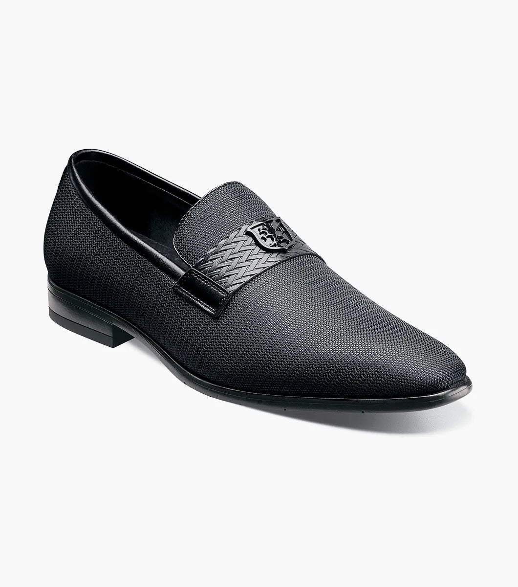 The STACY ADAMS Tazzi Plain Toe Saddle Ornament Slip On (25529-001) is a black textured men’s loafer with leather linings, Memory Foam footbed, decorative saddle band and emblem, and low heel—shown on a plain white background.