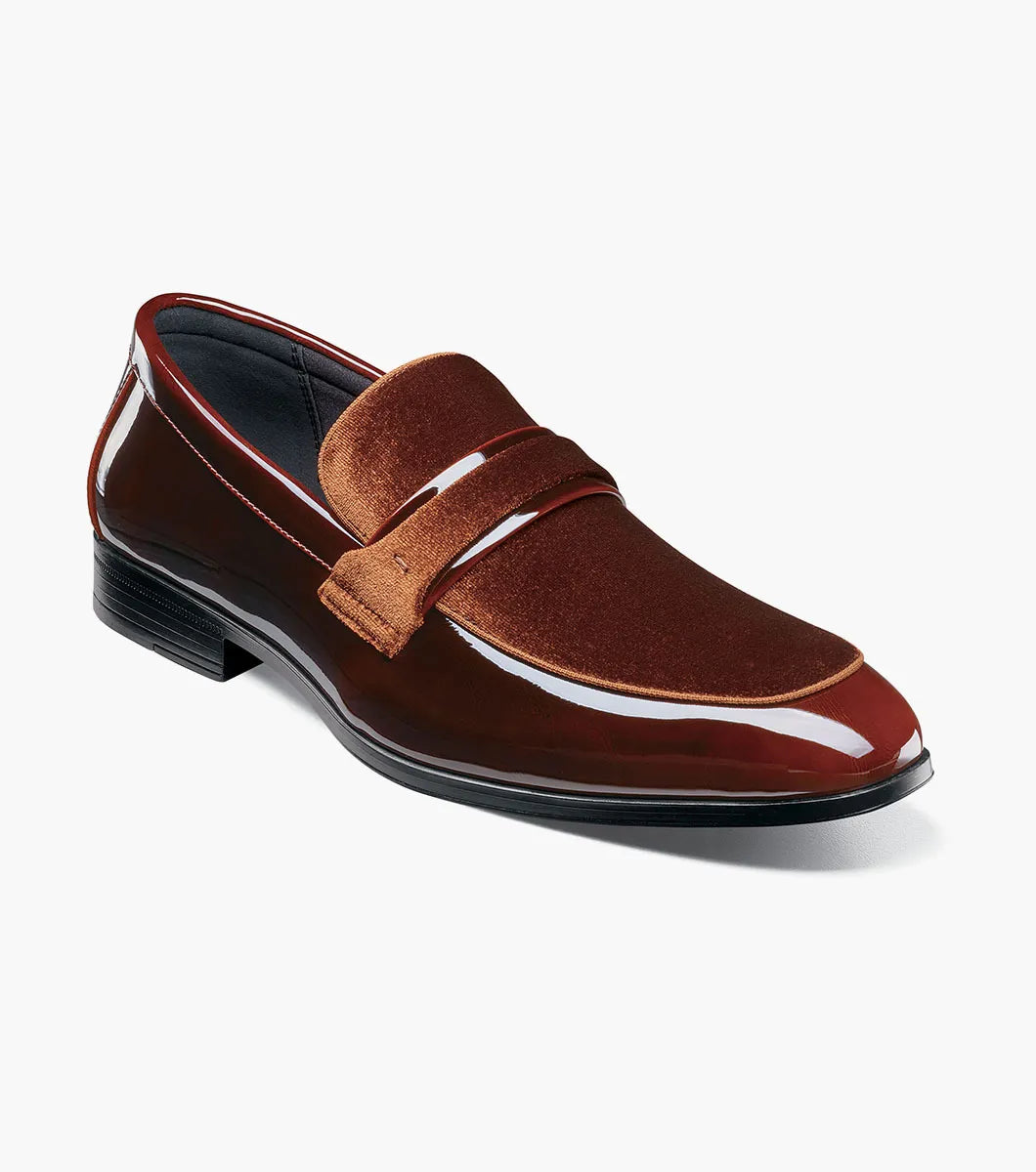 The Stacy Adams Spratley Moc Toe Saddle Slip On (Brown, 25686-200) is a men’s loafer with a glossy leather finish, cushioned insole, and velvet strap detail, displayed angled on a plain white background.