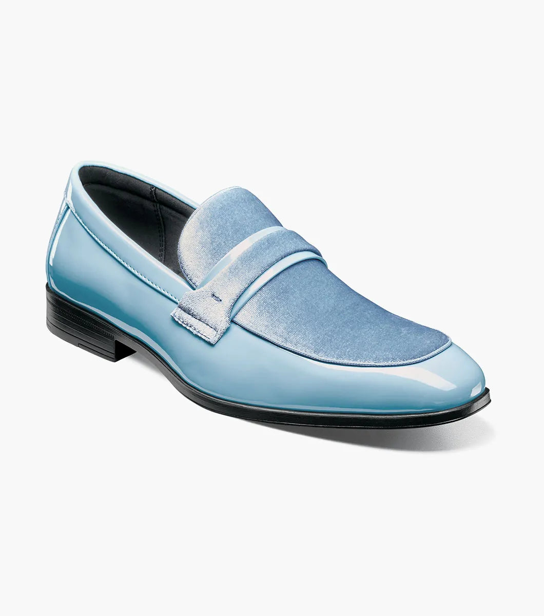 The Stacy Adams Spratley Moc Toe Saddle Slip On in light blue (style 25686-459) showcases a glossy patent leather body with a velour upper, complemented by a matching suede strap and vamp. This men’s loafer by STACY ADAMS features a cushioned insole for enhanced comfort, a black sole, and a low heel, all presented against a plain white background.