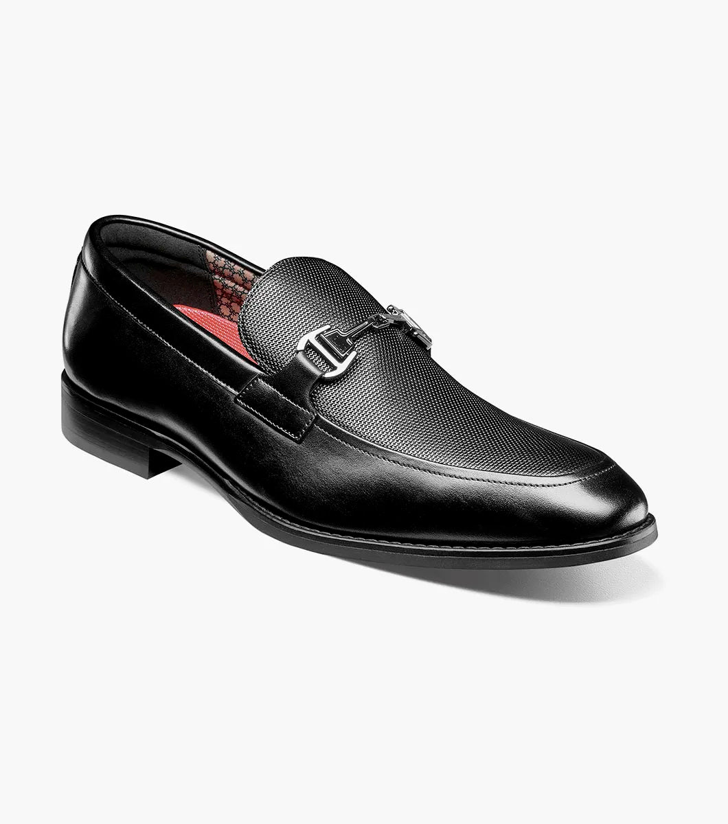 The Stacy Adams Lawton Moc Toe Bit Slip On in black features a textured leather upper, decorative metal buckle, and a classic moc toe slip-on design. This dress shoe by STACY ADAMS is finished with a low heel, red inner lining, and a cushioned footbed for added comfort. Product displayed on a white background.