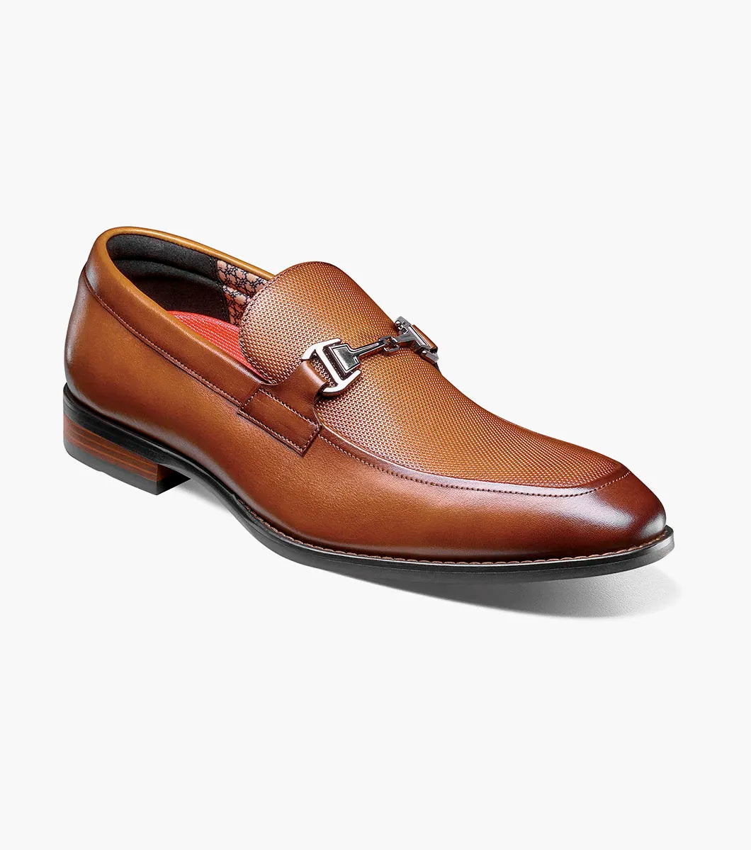 The Stacy Adams Lawton Moc Toe Bit Slip On in Cognac (style 25796-221) features a textured brown leather upper, classic moc toe slip-on design, cushioned footbed, silver metal bit detail across the vamp, and a low stacked heel. Shown at an angle on a white background, this STACY ADAMS shoe combines comfort with timeless style.