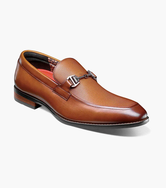 The Stacy Adams Lawton Moc Toe Bit Slip On in Cognac (style 25796-221) features a textured brown leather upper, classic moc toe slip-on design, cushioned footbed, silver metal bit detail across the vamp, and a low stacked heel. Shown at an angle on a white background, this STACY ADAMS shoe combines comfort with timeless style.