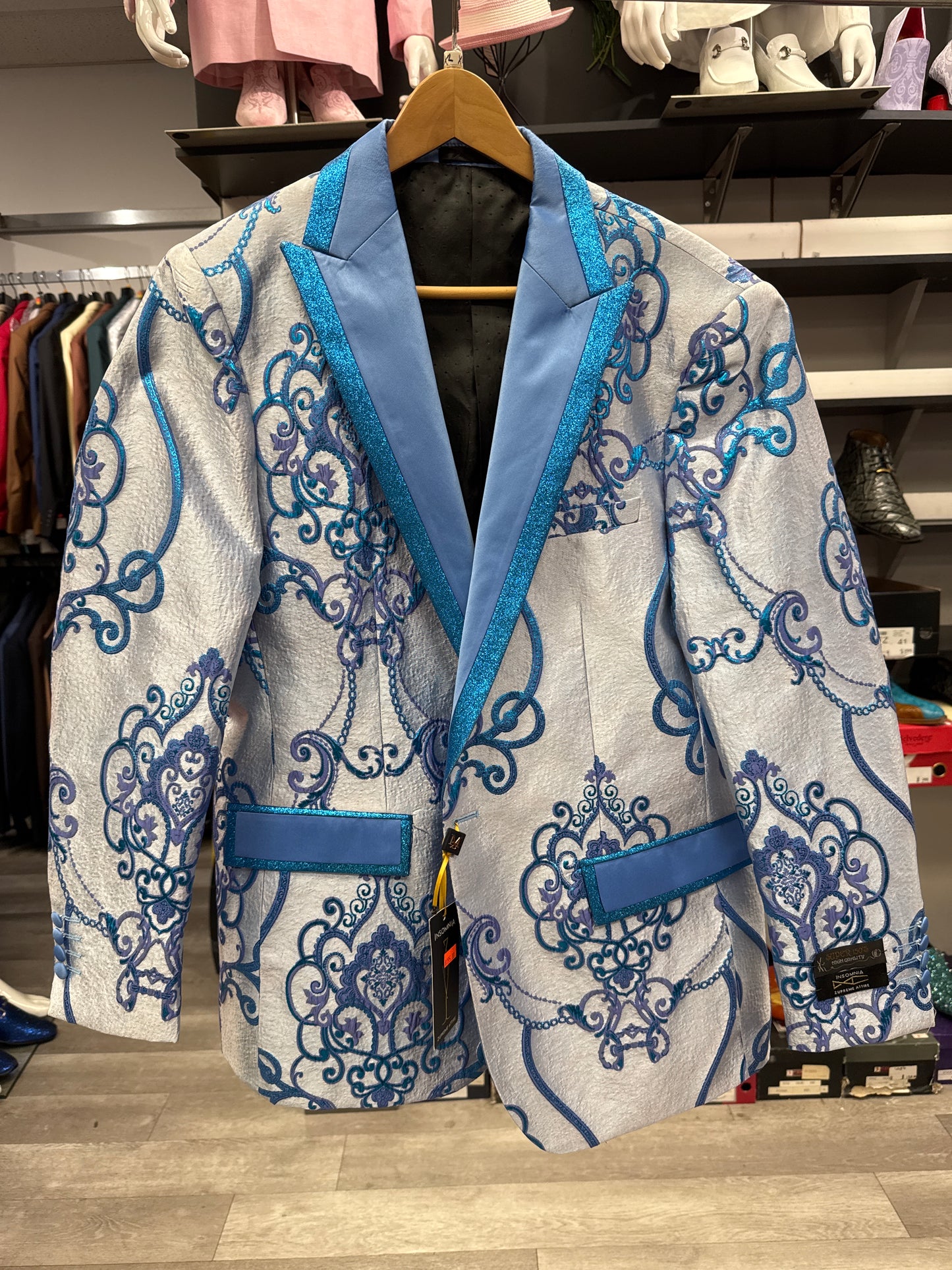 A shiny silver EJ SAMUEL JACKET 6 CLEARANCE, featuring blue ornate patterns, blue lapels, and blue-trimmed pockets, hangs on a wooden hanger in a clothing store—please note, this item is final sale.