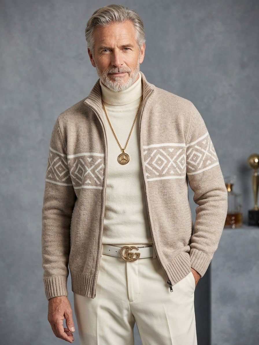 A man with gray hair and a beard wears the Suslo Couture Full Zip Sweater SW550 by Unique Design Menswear—a beige patterned full-zip cardigan—over a white turtleneck, paired with white pants, a white Gucci belt, and a gold necklace, standing against a gray background.