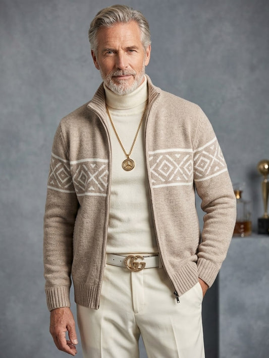A man with gray hair and a beard wears the Suslo Couture Full Zip Sweater SW550 by Unique Design Menswear—a beige patterned full-zip cardigan—over a white turtleneck, paired with white pants, a white Gucci belt, and a gold necklace, standing against a gray background.