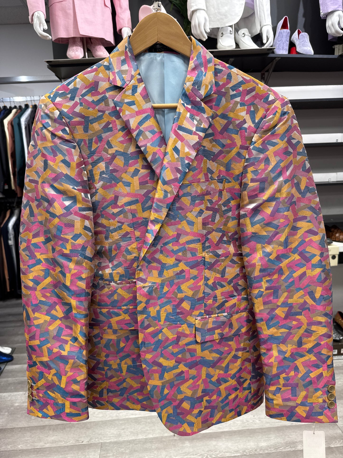 The EJ SAMUEL JACKET 24 CLEARANCE, a multicolored blazer featuring an abstract geometric pattern in pink, yellow, blue, and purple, hangs on a wooden hanger in a clothing store—please note this is a final sale item and is not eligible for exchanges or returns.