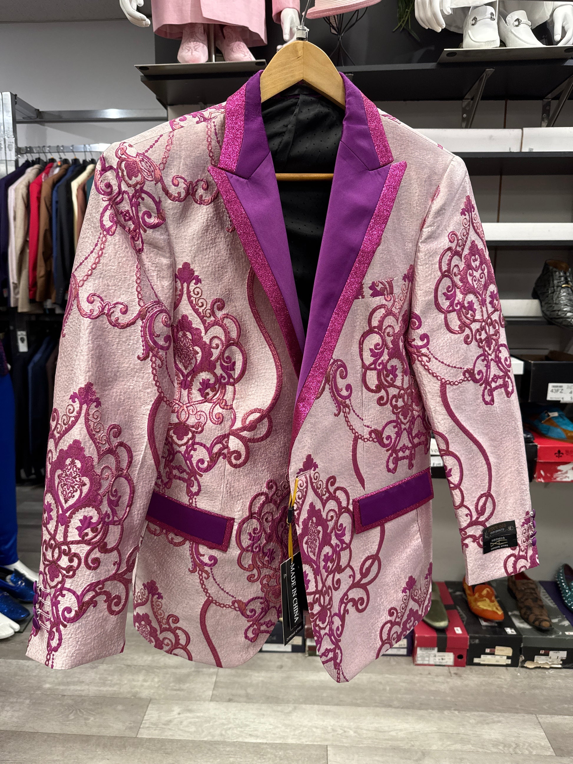 The EJ SAMUEL JACKET 7 CLEARANCE, featuring pink fabric with purple lapels and ornate magenta embroidery, hangs on a wooden hanger in a clothing store—please note, this is a final sale item with no exchanges or returns.