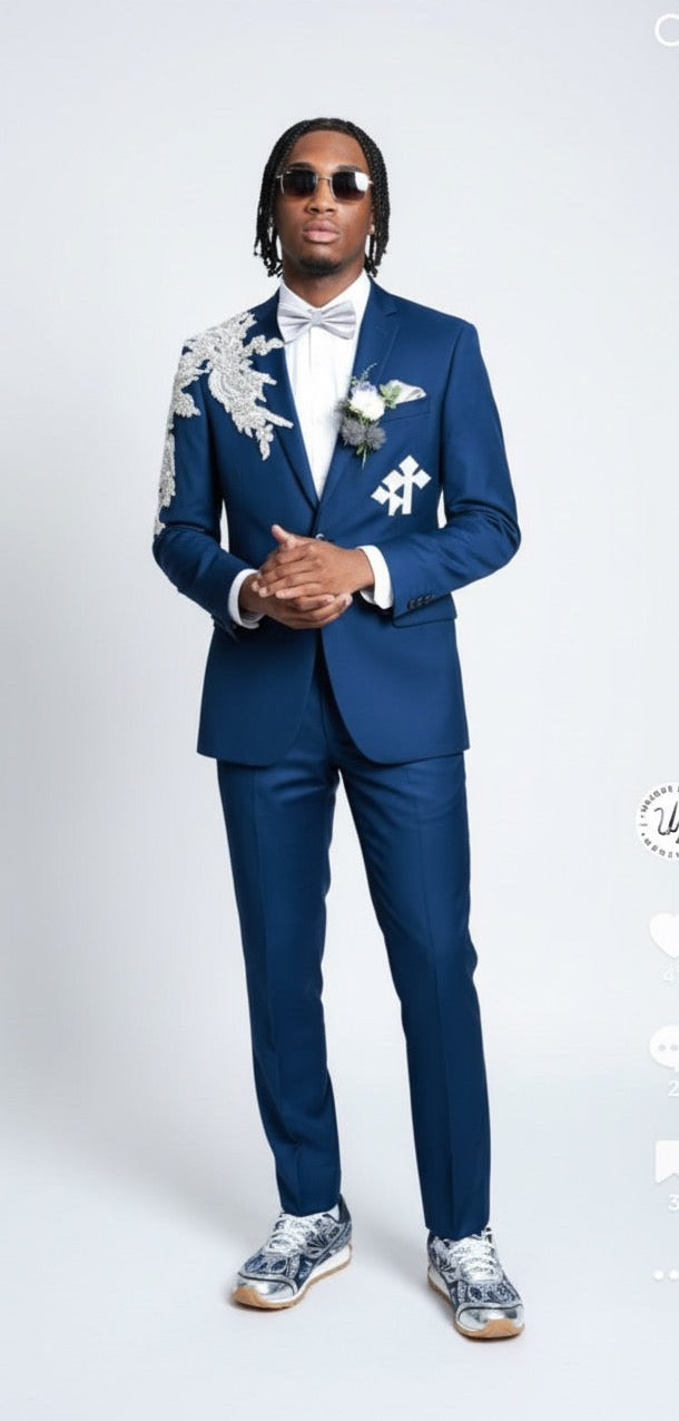 A man stands against a plain background wearing the Unique Design Menswear Custom Prom Suit in blue with silver rhinestones, paired with a white bow tie, sunglasses, and patterned sneakers.