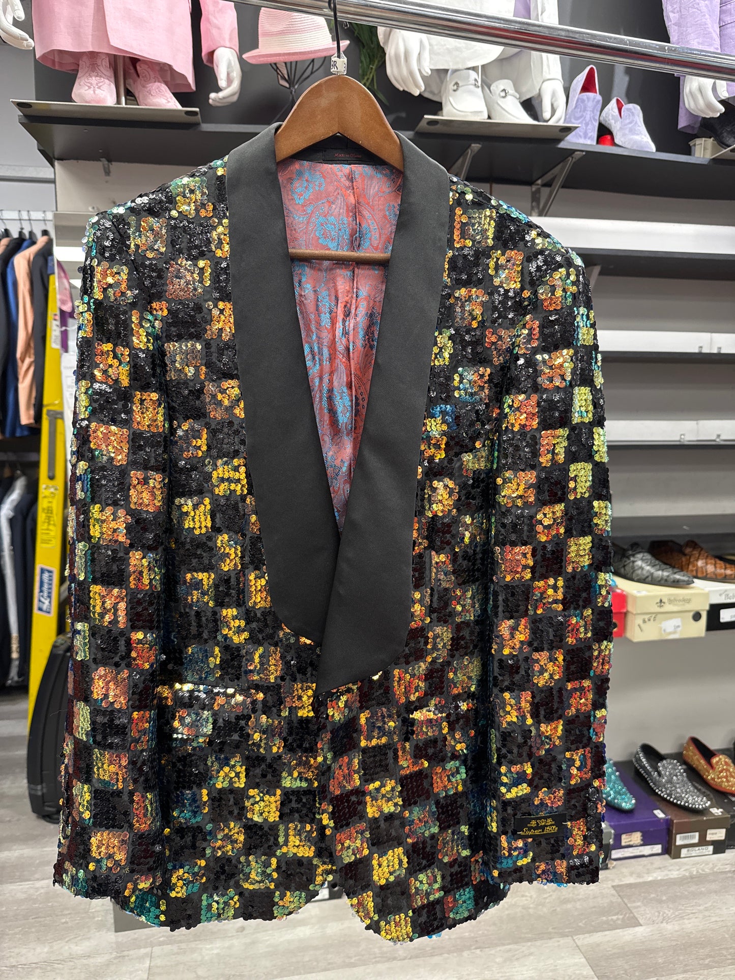 The EJ SAMUEL JACKET 2 CLEARANCE—a sequined blazer featuring a black shawl collar—hangs on a wooden hanger in a clothing store, surrounded by shirts, shoes, and other accessories. Please note: this item is final sale—no returns or exchanges allowed.