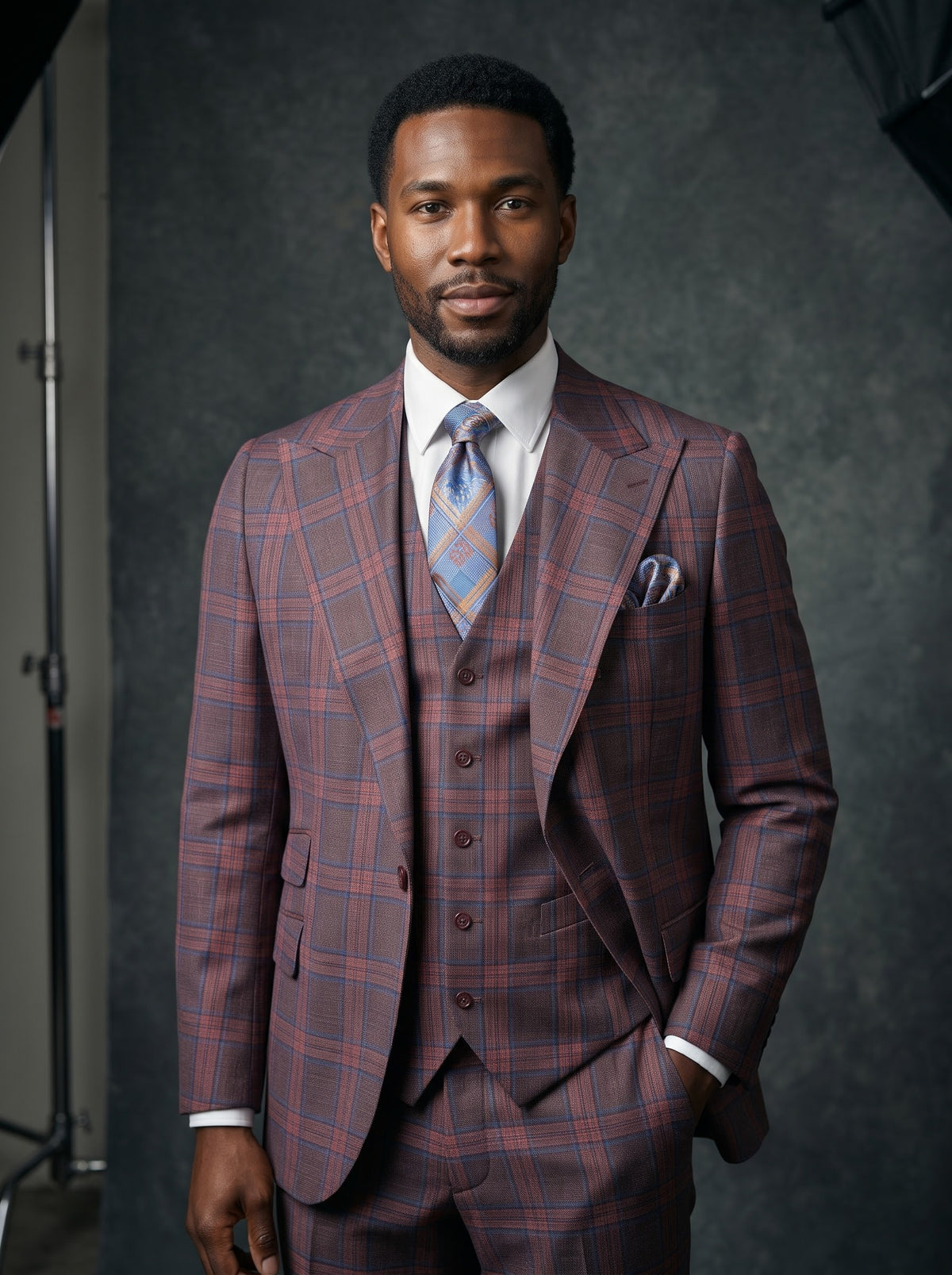 A man wears a Tiglio Luxe Antibes Brick/Blue Plaid Semi Wide Leg Suit TL8305 featuring a brick-red and blue plaid three-piece design, white dress shirt, patterned blue tie, and matching pocket square.