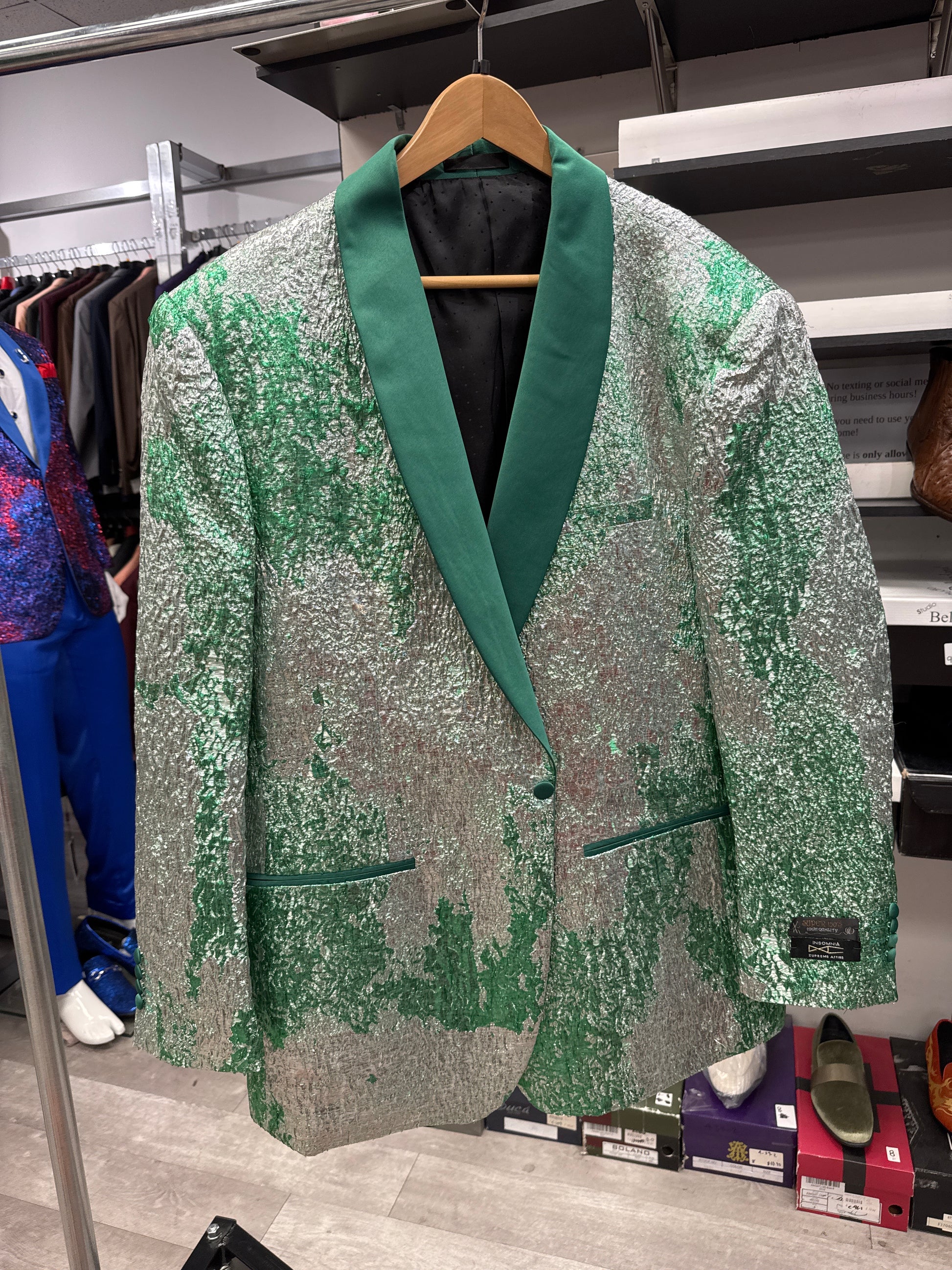 The EJ SAMUEL JACKET 9 CLEARANCE, a silver and green sequin blazer featuring a dark green shawl collar, is displayed on a wooden hanger in a clothing store among other garments and shoes. Please note: this item is final sale with no exchanges or returns.