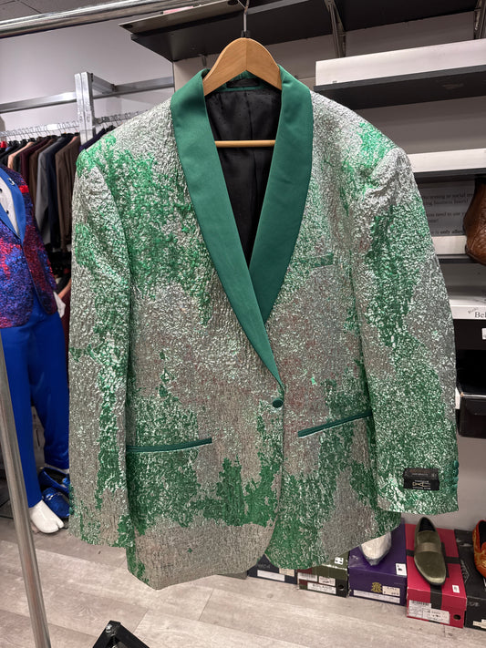 The EJ SAMUEL JACKET 9 CLEARANCE, a silver and green sequin blazer featuring a dark green shawl collar, is displayed on a wooden hanger in a clothing store among other garments and shoes. Please note: this item is final sale with no exchanges or returns.