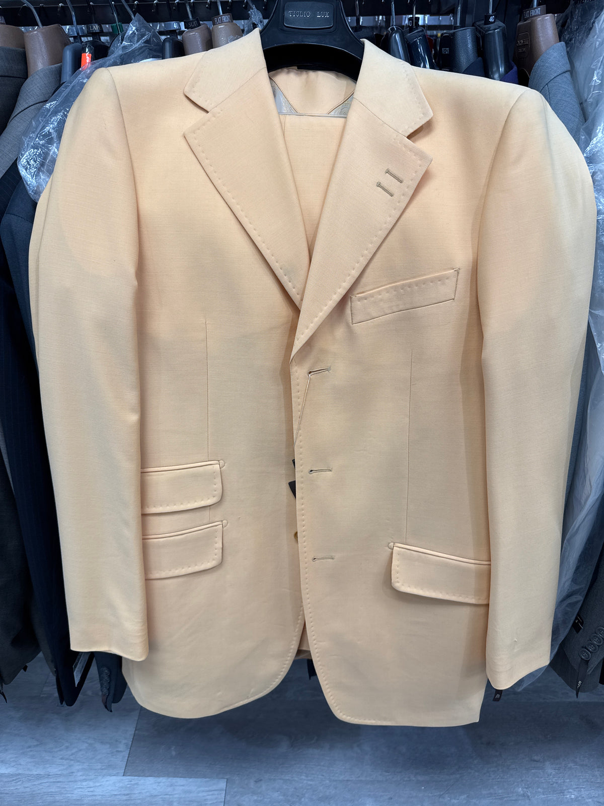A man stands indoors smiling, dressed in the Tiglio Rosso “BASITO” Banana Wide Leg Pure Wool Suit (E09063-1), paired with a white shirt, tie, and brown shoes. A potted plant and beige backdrop are visible in the background.