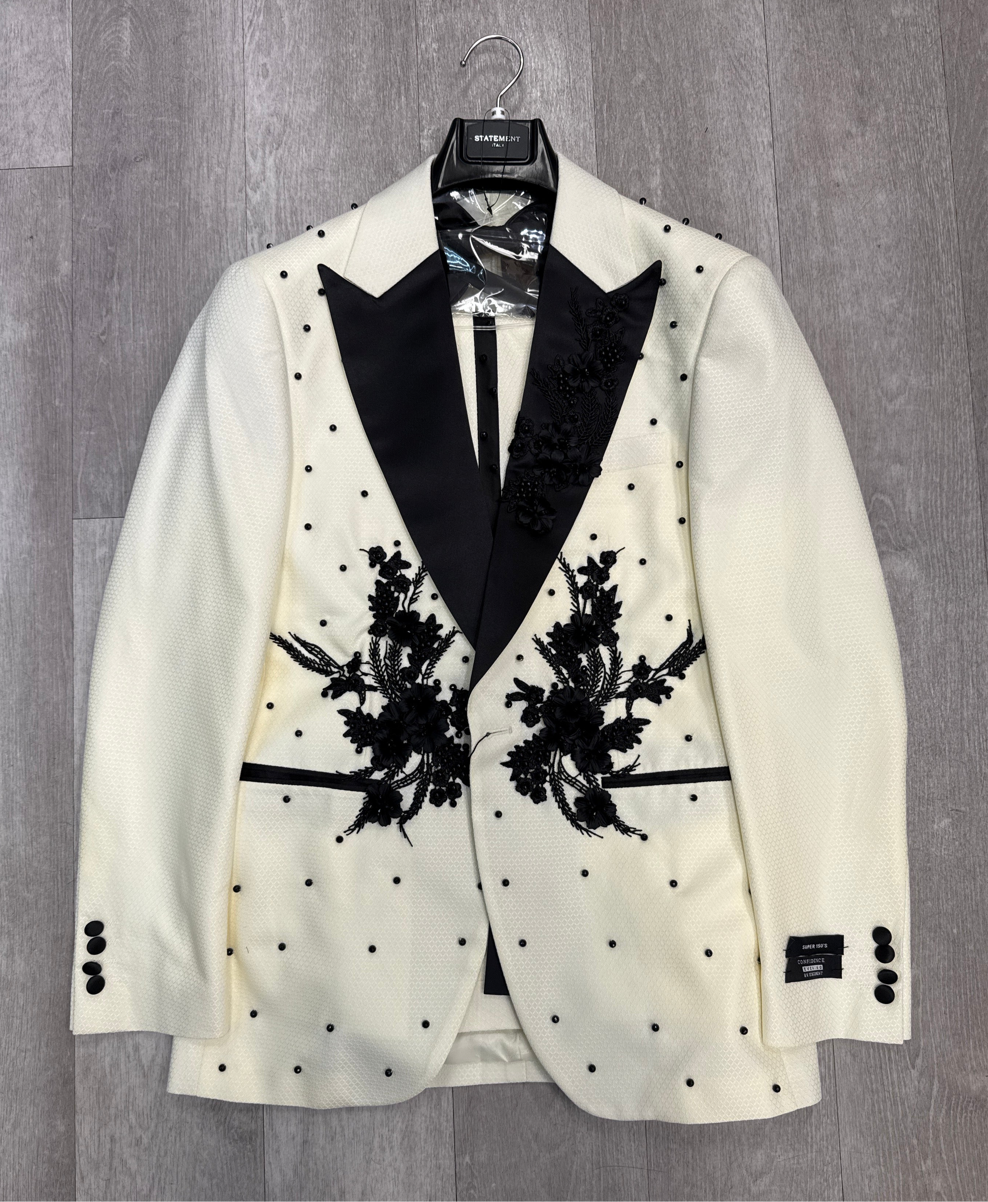The STATEMENT CLOTHING | PERINI - OFFWHITE - BLACK tuxedo jacket by Statement Clothing features a cream color, black lapels, black floral embroidery, and black polka dot accents, shown on a hanger against a gray wood floor.